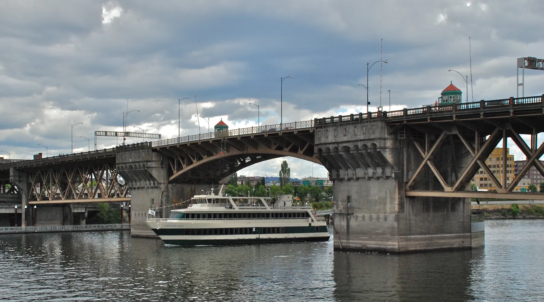 En turistbåd, der passerer under Burnside Bridge, med betonpillerne og stålbuerne over en grågrøn flod