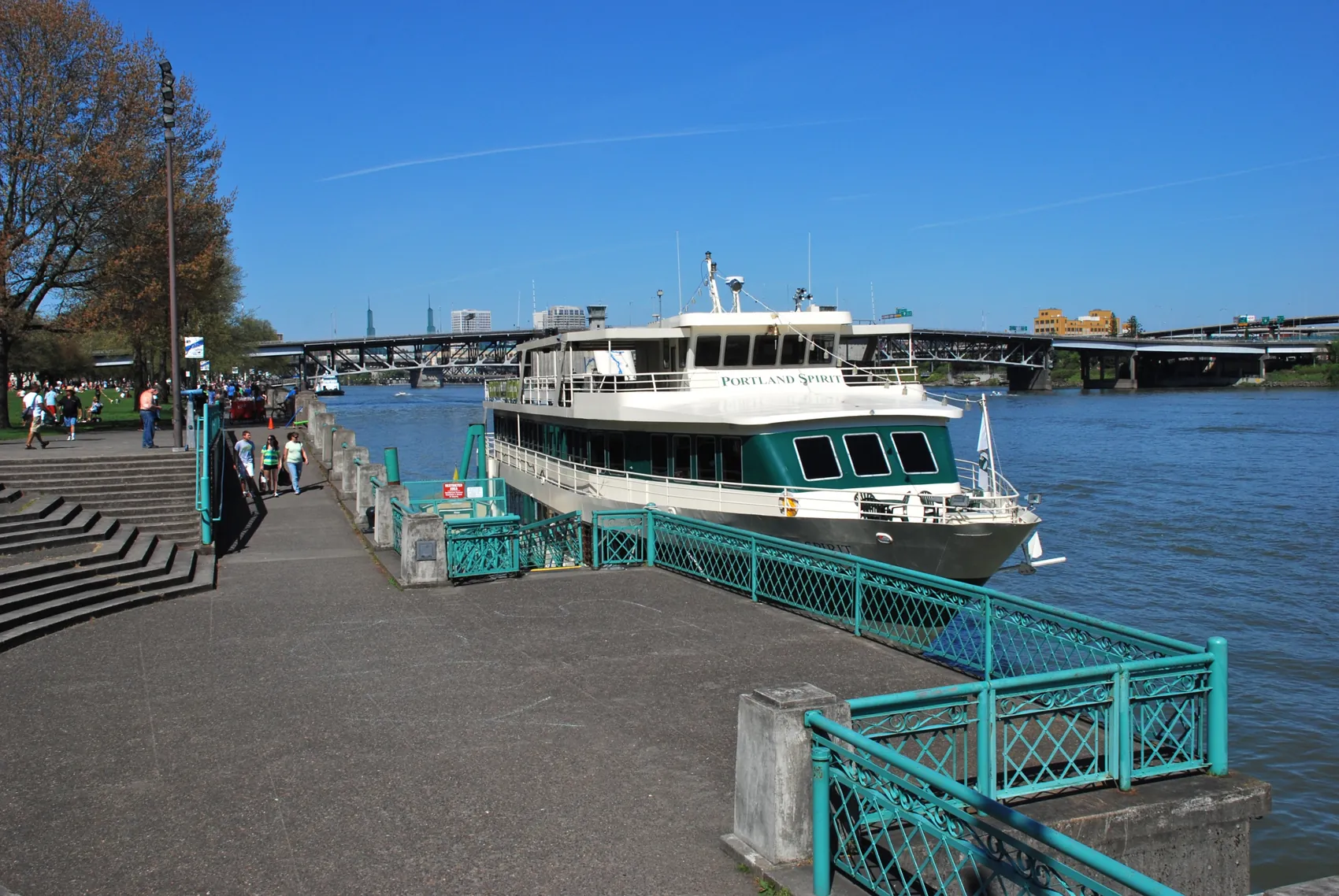 En Portland Spirit-båd fortøjet ved Waterfront Park-broen ved siden af betontrin og et blågrønt rækværk under en blå himmel