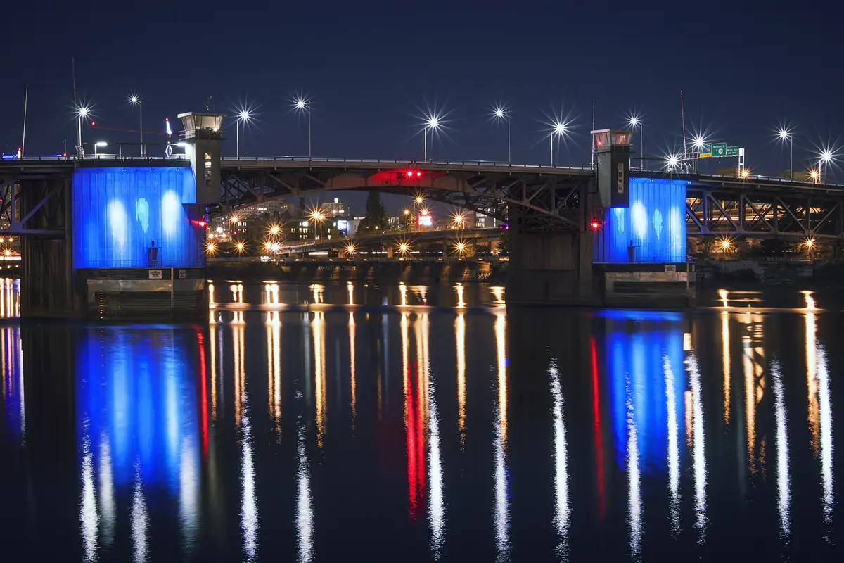 A Morrison Bridge à noite, com os seus dois pilares de betão iluminados a azul e a faixa rodoviária ladeada de lâmpadas brilhantes, refletidos no rio