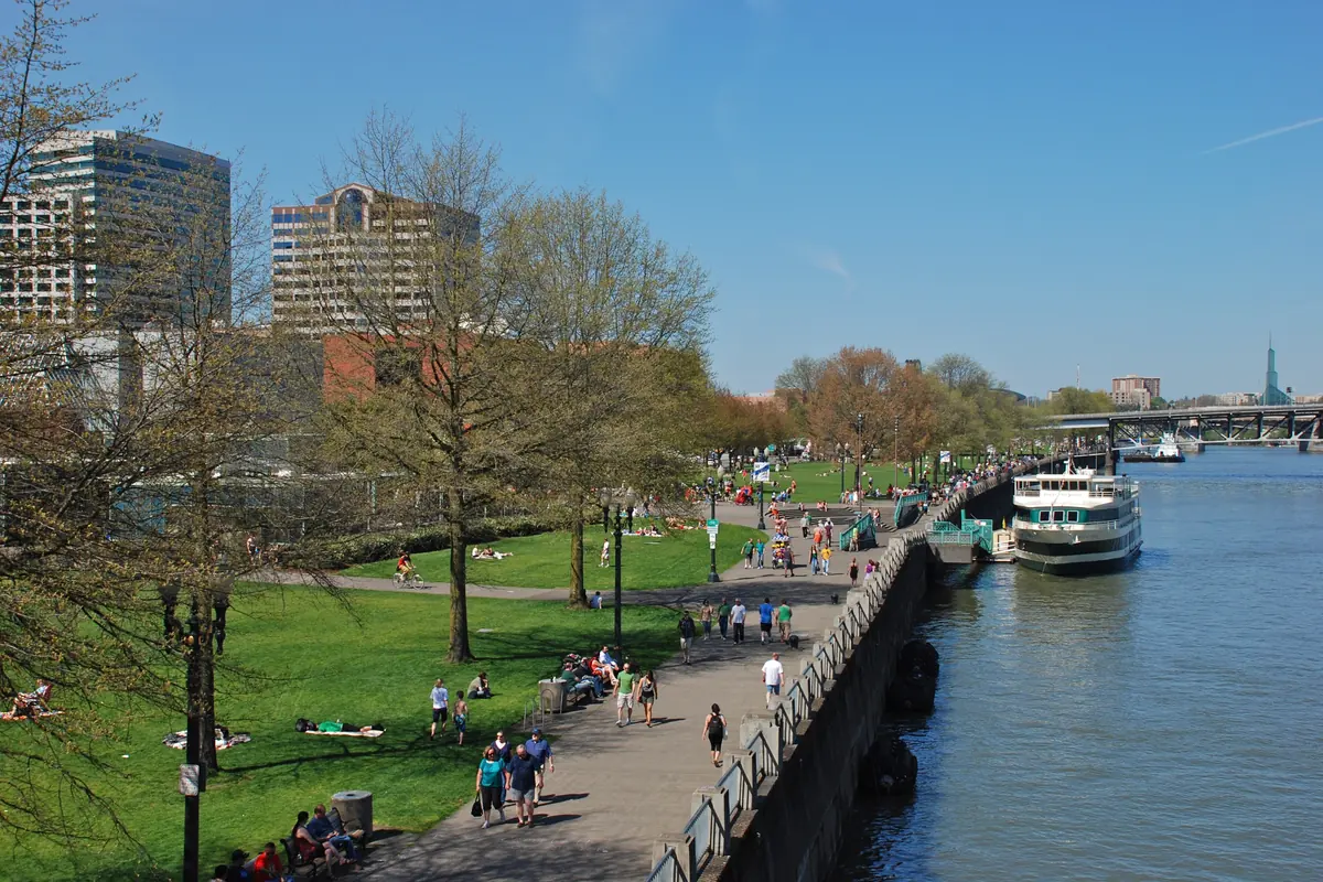 Spacerowicze i osoby urządzające pikniki na trawnikach i ścieżce wzdłuż muru nadbrzeżnego w Tom McCall Waterfront Park, z łodzią wycieczkową zacumowaną przy pomoście